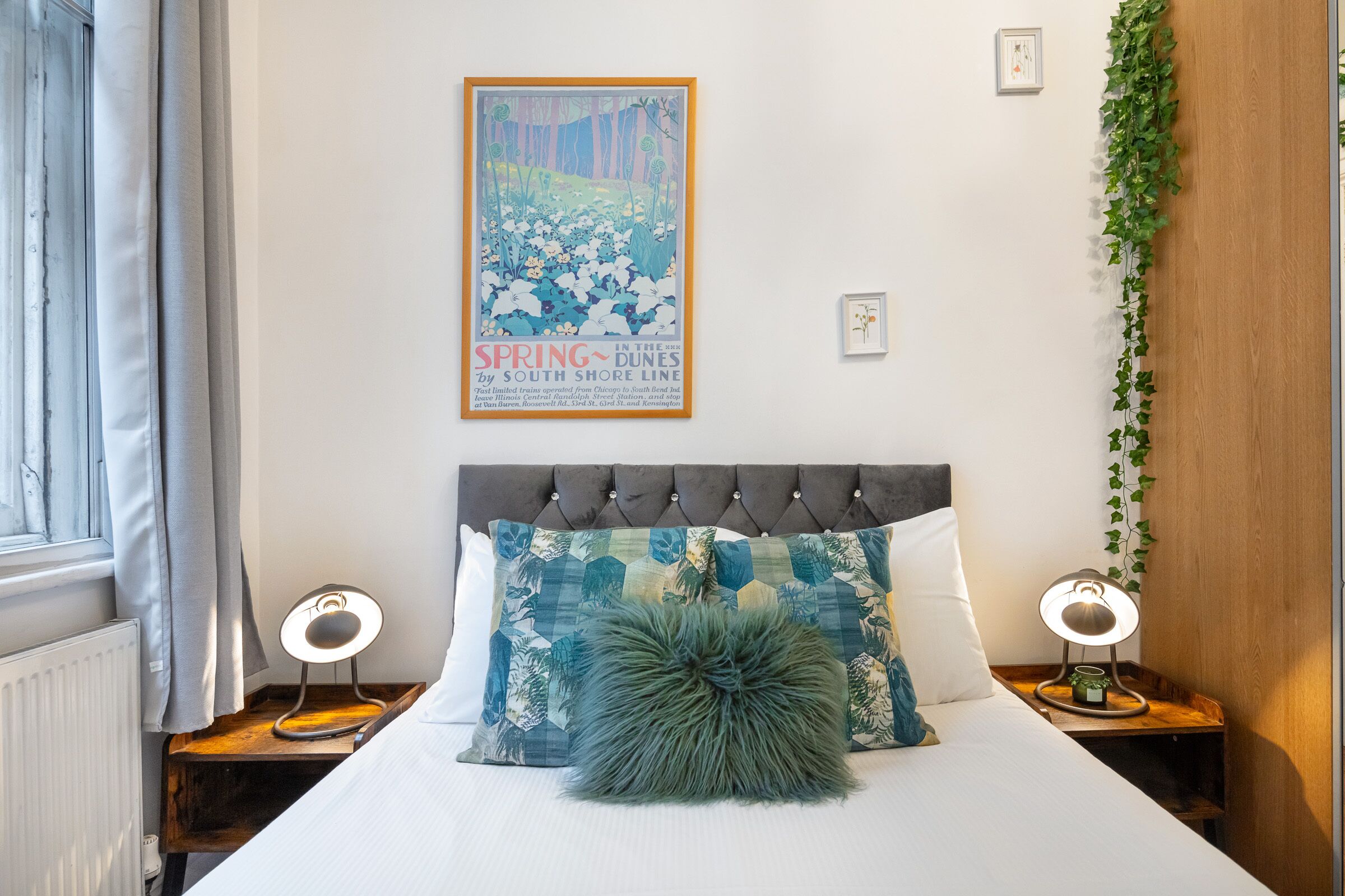 Un dormitorio con una cama, dos mesitas de noche con lámparas, un cartel en la pared y una ventana con cortinas.