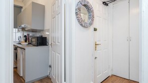 A compact kitchen with white cabinets, a microwave, and a sink. A white door with a gold handle and a circular mirror with magazine pages are visible.