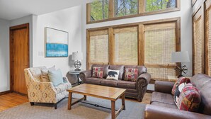 A living room with a brown leather sofa, a patterned armchair, and a wooden coffee table. There are two windows with wooden blinds, a painting on the wall, and a lamp on a side table. - Ober Gatlinburg | Private Hot Tub | Arcade | Free High-Speed Wi-Fi (Gatlinburg)