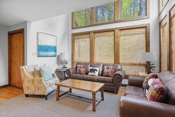 A living room with a brown leather sofa, patterned armchair, and a wooden coffee table. There are two windows with wooden frames and blinds, and a painting on the wall.