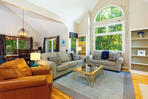 A spacious living room with a high ceiling, a large window with a view of trees, a leather sofa, a glass coffee table, and a bookshelf.