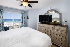 A bedroom with a large bed, a dresser, a television, a ceiling fan, and a view of the beach.