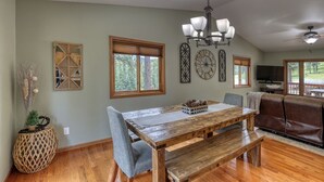 A dining area with a wooden table, benches, and a chandelier. There are decorative wall pieces and a large window with blinds. - Sunny Day Hideaway - Private! Firepit and Hot tub! (Sturgis)