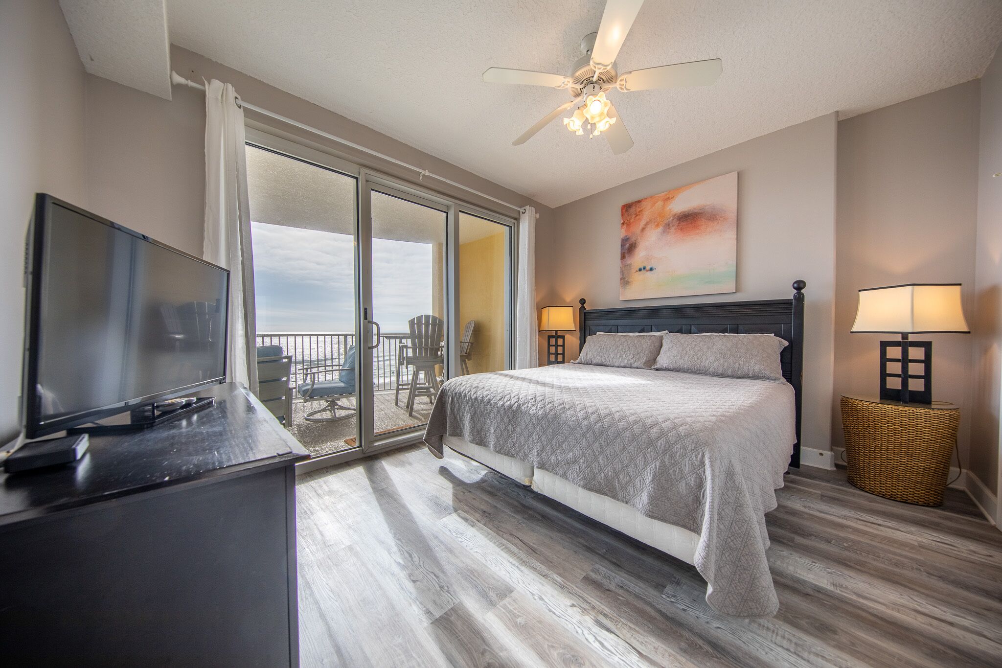 A bedroom with a bed, a television, a ceiling fan, and a balcony with chairs.