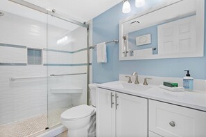 A bathroom with a glass shower enclosure, a white toilet, a sink with a mirror above, and a blue wall.