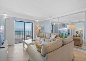 A modern living room with a large sofa, a coffee table, and a TV. There is a balcony with a view of the ocean.