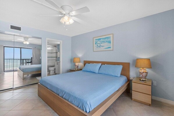 A bedroom with a bed, bedside tables, a ceiling fan, a painting, and a view through a glass door.