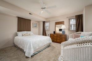 Un dormitorio con cama, cómoda, silla de mimbre, ventilador de techo y ventana con cortinas.