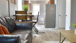Una sala de estar con un sofá de cuero negro, una mesa de centro de madera, un tapete estampado, una mesa de comedor con sillas, un refrigerador y una puerta.
