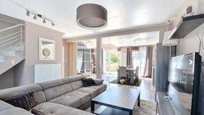 A modern living room with a grey sofa, a coffee table, and a flat-screen TV. There is a staircase on the left and a dining area with a table and chairs in the background. - House 120 m² in Vigy (Vigy)