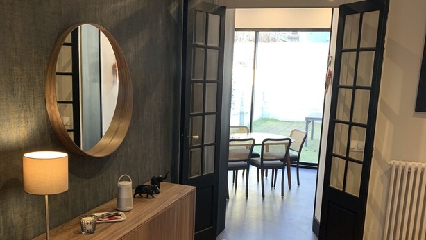 A room with a wooden console table, a round mirror, and a table lamp. There is a door leading to an outdoor area with a table and chairs.