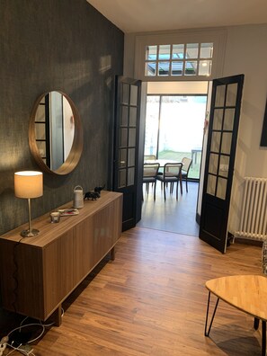 Una habitación con una consola de madera, un espejo redondo y una lámpara de mesa. Hay una puerta que da a un espacio exterior con una mesa y sillas.