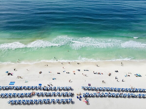A beach with rows of blue and white umbrellas, people sunbathing and swimming, and waves crashing onto the shore.