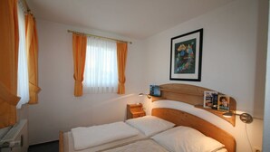 Quarto com uma cama de solteiro, mesa de cabeceira, estante com livros, quadro na parede e janela com cortinas.