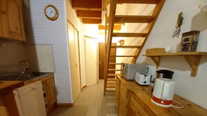 Eine kleine Küche mit Holzausbau, Mikrowelle, Toaster und Kaffeemaschine auf einer Holz Arbeitsplatte. Eine Uhr ist an der Wand sichtbar, und eine Holzleiter führt zu einem Erkerraum.
