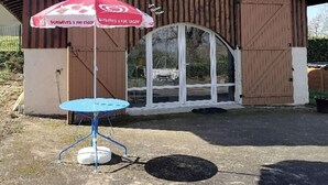 A rustic wooden building with a large arched entrance, a small table with a blue top, and a red umbrella with white text. - Barn converted into gîte for 4/5 people (audon)