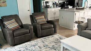 Un salon moderne avec deux fauteuils en cuir, une table basse blanche et un canapé gris. On aperçoit, au fond, une cuisine avec des meubles blancs et un réfrigérateur noir.