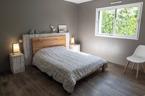 A bedroom with a wooden headboard, a bed with patterned bedding, bedside lamps, a window with a view of greenery, and a white chair.