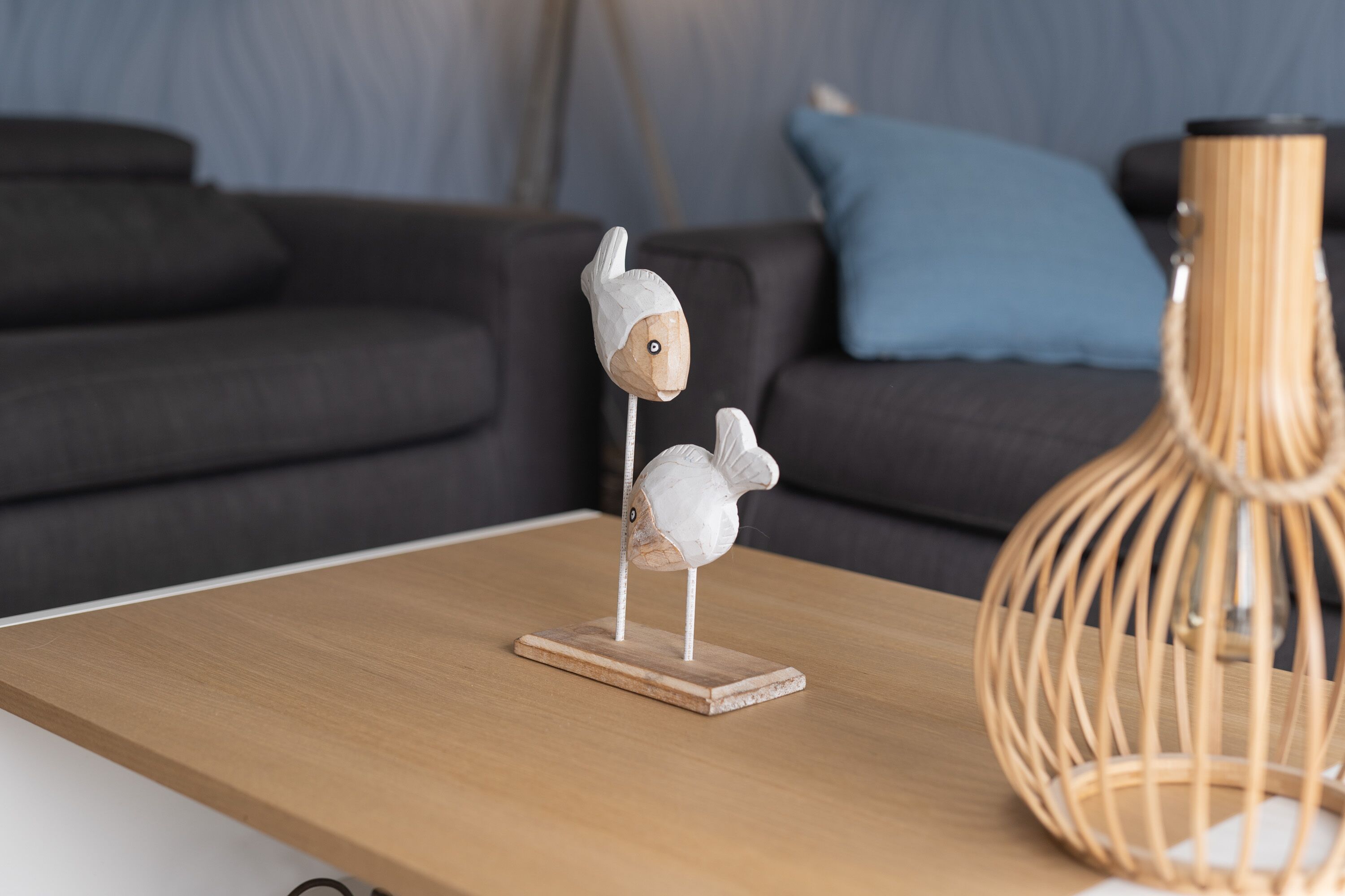 A wooden table with two white bird sculptures on stands, a woven lamp, and a dark sofa with blue cushions in the background.