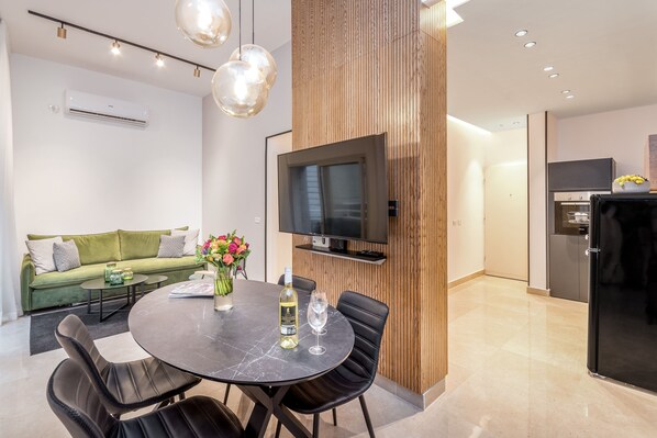Ein modernes Wohnzimmer mit einem grünen Sofa, einem Esstisch mit Stühlen, einem an der Wand montierten Fernseher und einer sichtbaren Küchenzeile im Hintergrund.