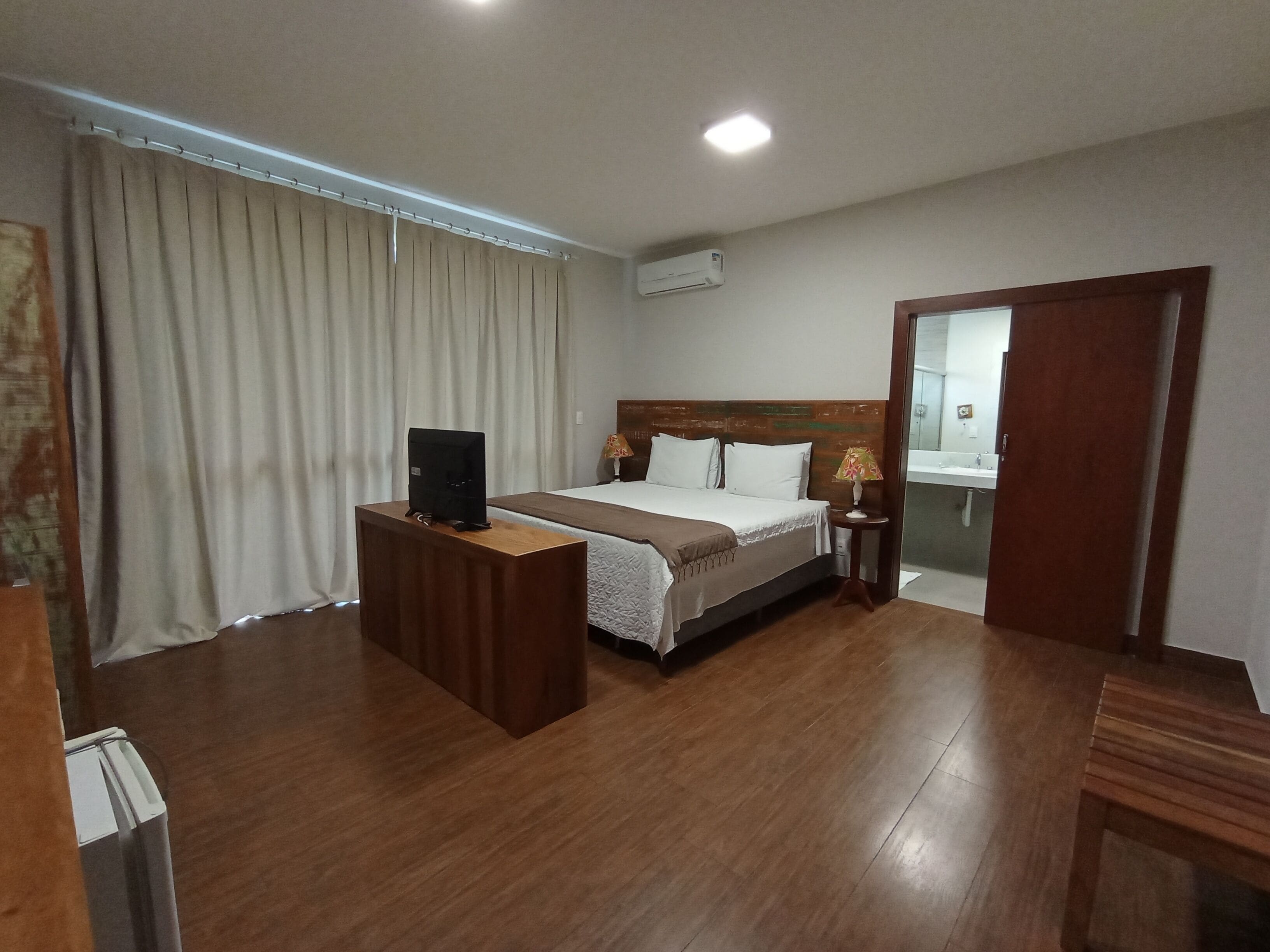 Ein Hotelzimmer mit Holzboden, einem Bett mit weißer Bettwäsche, einem Fernseher auf einem Holztischchen, einem Nachttisch mit Lampe und einem durchgehenden Türspalt einsehbarer Badezimmers.