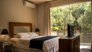 A bedroom with a bed, bedside tables, a TV on a wooden stand, and a large window with a view of trees. - Pousada Recanto das Araras (Tiradentes)