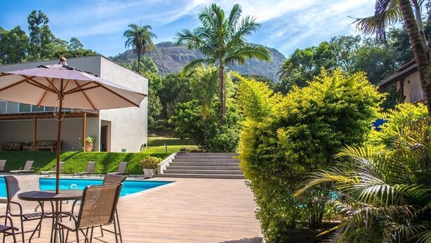 A pool area with lounge chairs and an umbrella, surrounded by lush greenery and a mountain backdrop. - Pousada Recanto das Araras (Tiradentes)