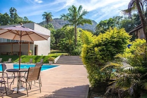 A pool area with lounge chairs and an umbrella, surrounded by lush greenery and a mountain backdrop. - Pousada Recanto das Araras (Tiradentes)