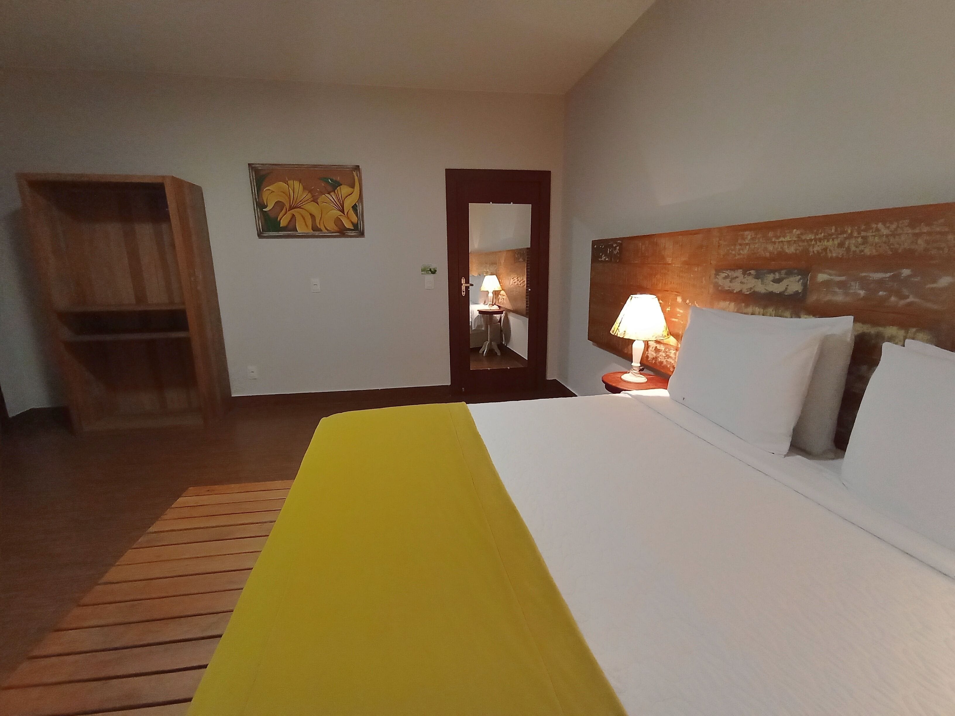 A hotel room with a bed, a wooden wardrobe, a painting on the wall, and a nightstand with a lamp.