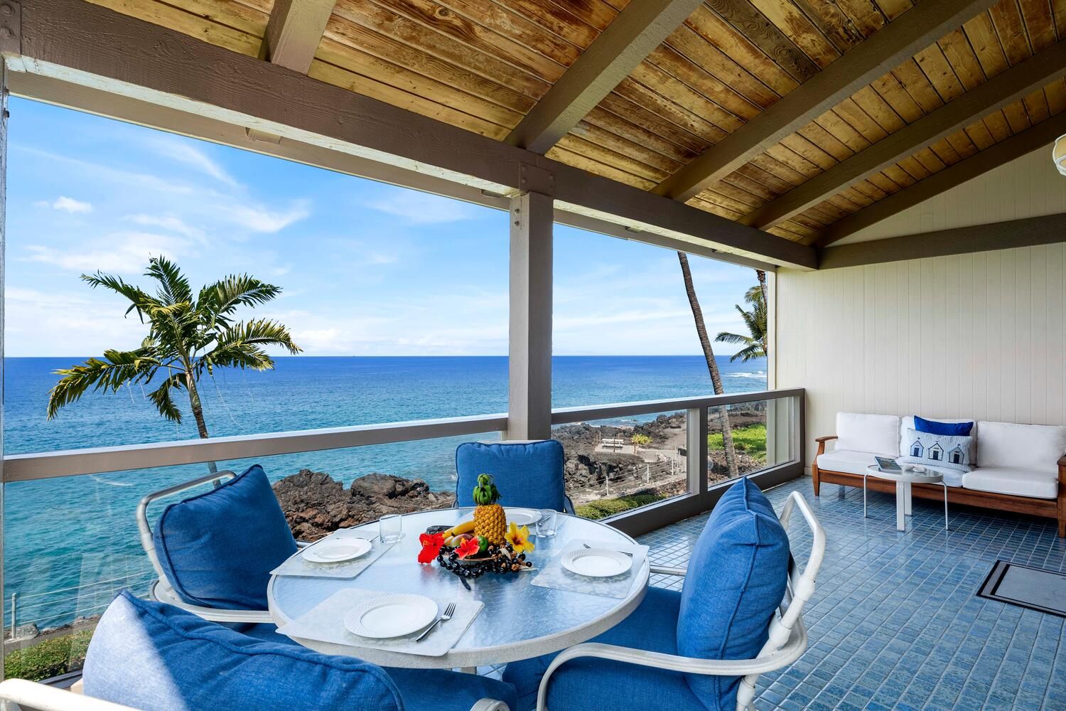 A dining area with a table set for two, overlooking the ocean with a view of the beach and palm trees.
