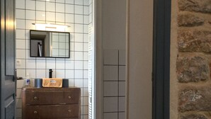 Un couloir étroit doté d’un plancher en bois, d’un mur carrelé et d’un mur en pierre sur la droite. On aperçoit au fond une commode en bois sur laquelle est posée une serviette.