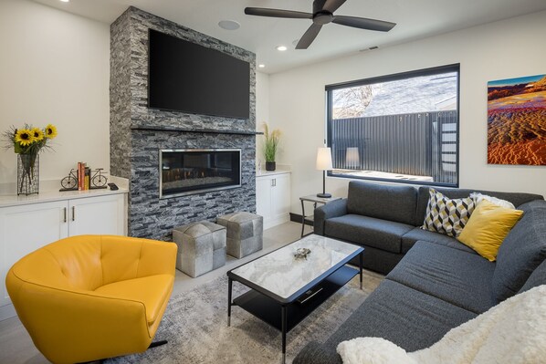 A modern living room with a stone fireplace, a flat-screen TV, a sofa, a yellow armchair, and a large window with a view. - Downtown Moab | Steps from Bike Path & Restaurants (Moab)