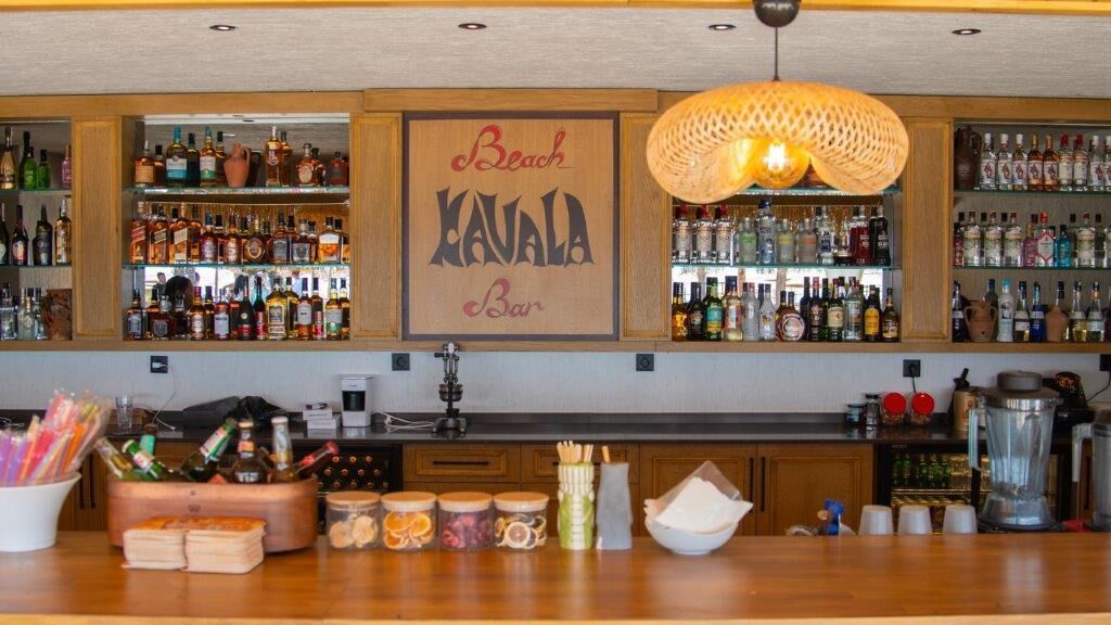 A bar with a wooden counter, shelves of liquor bottles, and a sign that reads "Beach Kavala Bar".