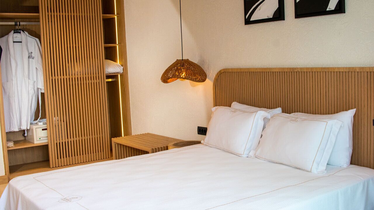 A hotel room with a bed, a bedside table, a hanging light fixture, a wooden wardrobe, and a wall-mounted artwork.