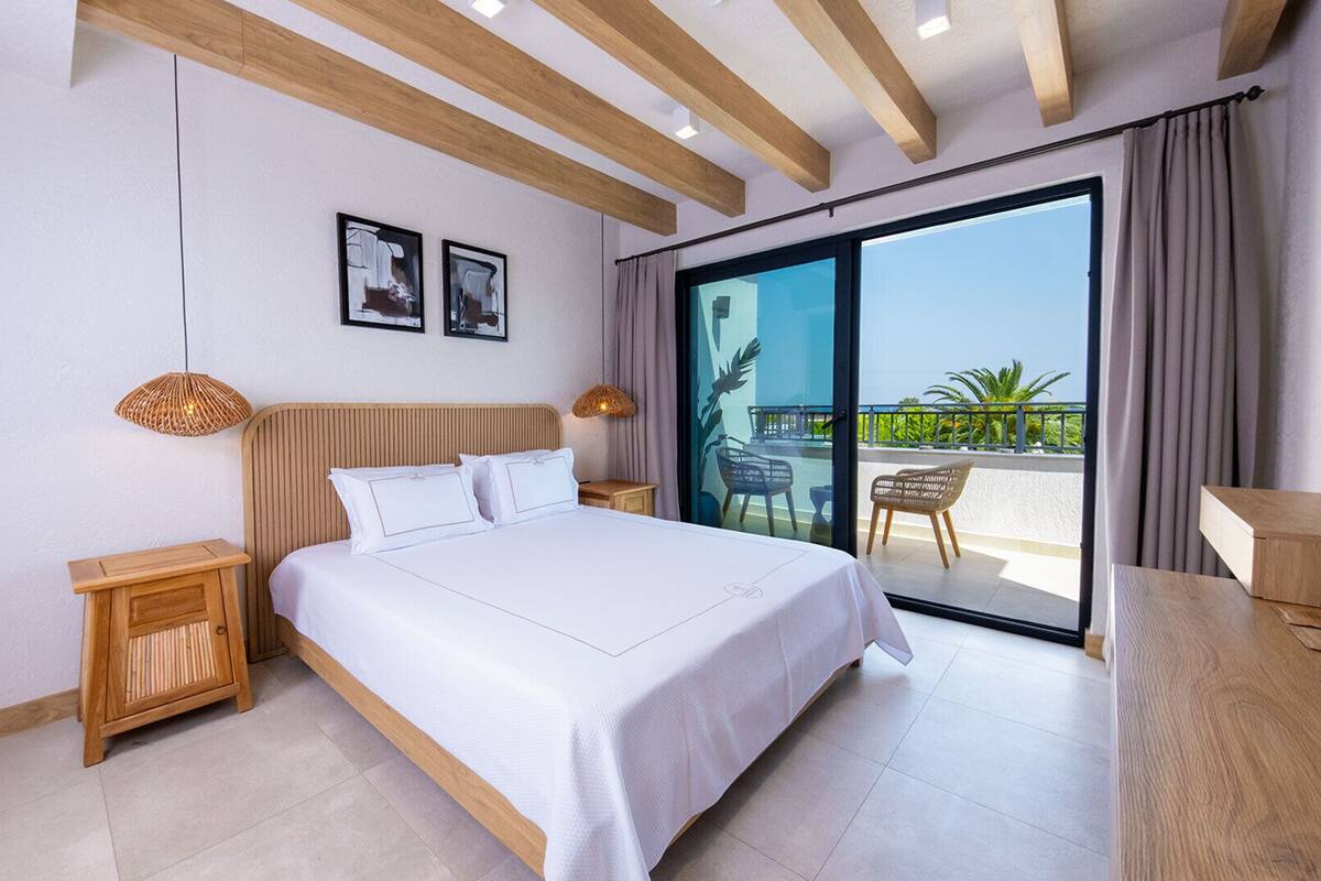 Une chambre d’hôtel moderne avec un grand lit, une tête de lit en bois, deux tableaux accrochés au mur, un balcon avec vue et une petite table d’appoint en bois.