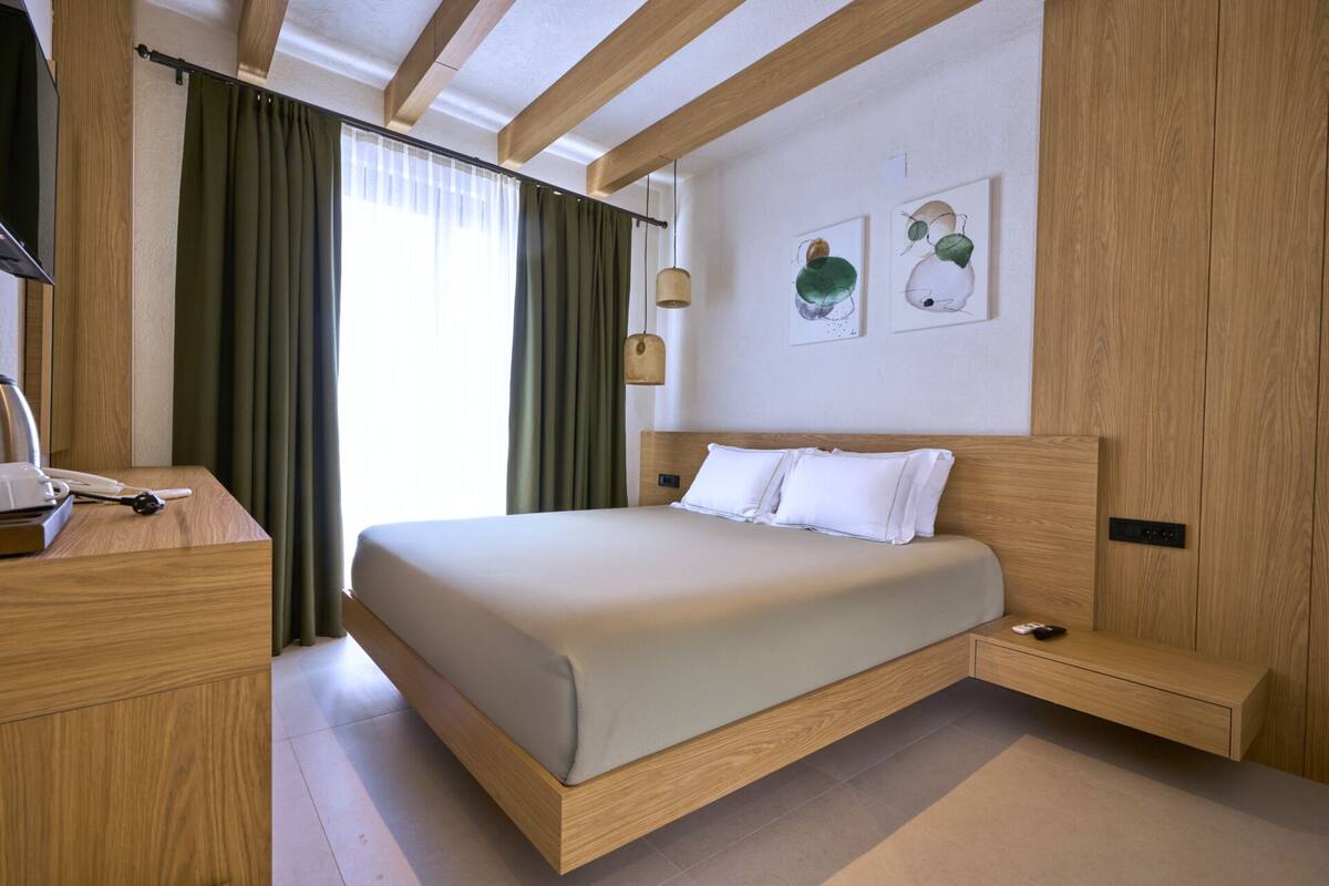 Une chambre d’hôtel moderne avec un cadre de lit en bois, une table de chevet et un bureau avec une lampe. Deux œuvres d’art sont accrochées au mur, et il y a une fenêtre avec des rideaux.