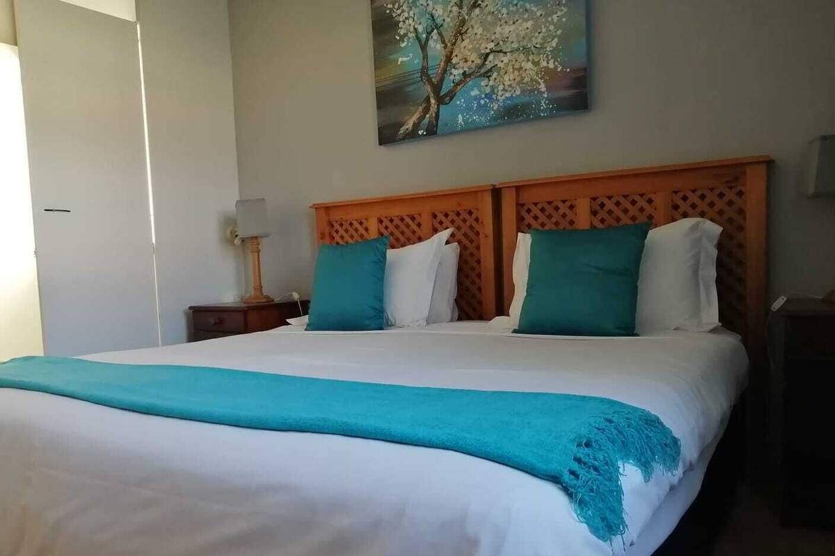 Una cama bien tendida con una colcha blanca y turquesa, un cabecero de madera y un cuadro de un árbol en la pared.