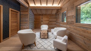 Interior de uma cabana de tronco com teto alto, paredes de madeira e uma janela grande. Há três poltronas brancas estofadas e uma mesa de centro de madeira.