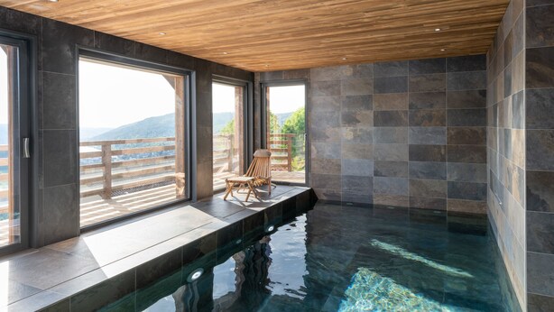 Piscina interior moderna com teto de madeira, janelas amplas e vista para o exterior.