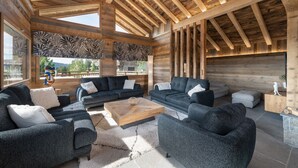 Uma sala de estar ampla, com sofás cinza escuro, uma mesa de centro de madeira e uma janela grande com vista para o exterior.