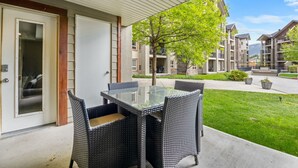 Une terrasse couverte avec une table en verre et des chaises en osier, une porte et une vue sur un ensemble d’immeubles résidentiels.
