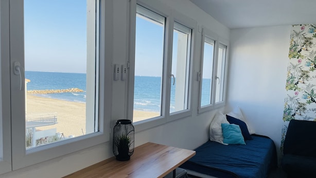 A bedroom with a wooden desk, a bed with blue bedding, and a view of the beach through large windows. - Waterfront studio, sea view 1st line beach (Palavas)