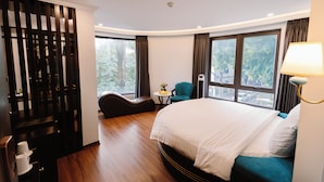 A hotel room with a large bed, a seating area with a chair and a small table, a decorative wall panel, and large windows with curtains. - Hồng Vận Hotel (Hanoi)