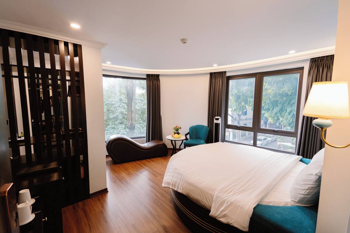 A hotel room with a large bed, a seating area with a chair and a small table, a decorative wall panel, and large windows with curtains.