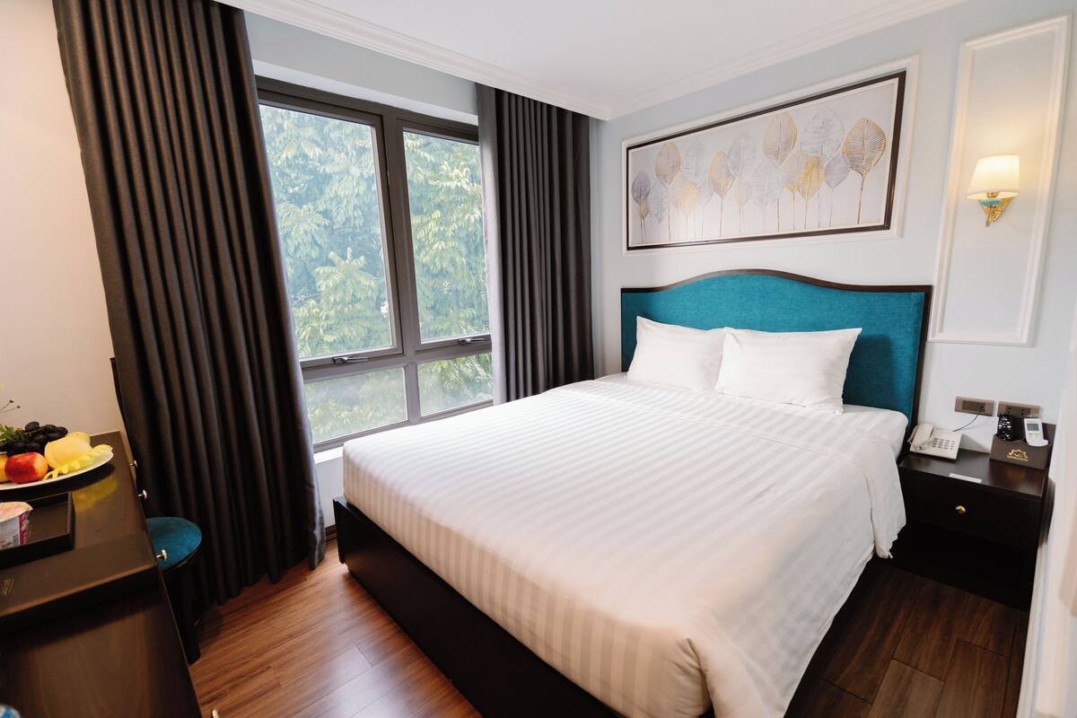 A hotel room with a large bed, a desk with a fruit bowl, a nightstand with a phone, and a window with curtains.