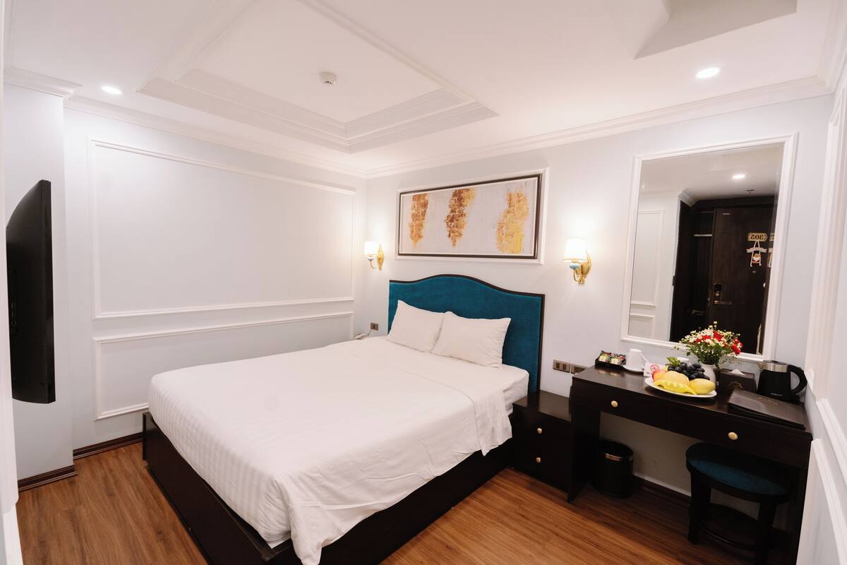 A hotel room with a large bed, a desk with a chair, a mirror, and a wall-mounted TV.