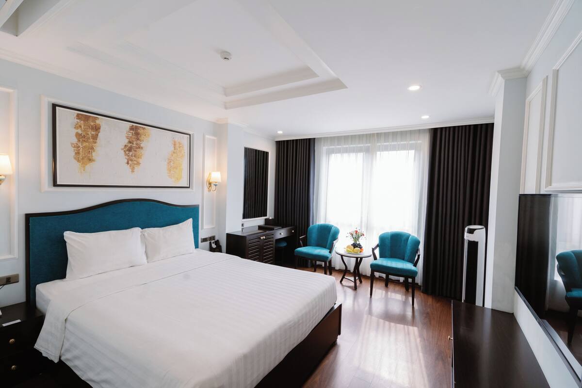 A hotel room with a large bed, two armchairs, a small table with a vase, and a TV.