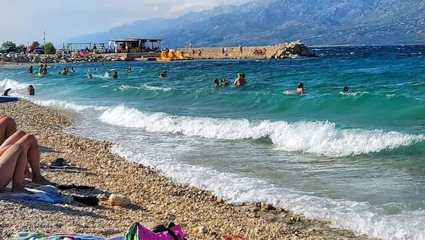 A beach with people swimming and sunbathing, a rocky shoreline, and mountains in the background. - Apartments Tara (Razanac)