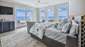 Une chambre dotée d’un grand lit, d’un ventilateur de plafond et d’une télévision. Elle offre une vue sur la plage grâce à de grandes fenêtres.