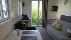 A compact room with a kitchenette, a dining table, a sofa with cushions, a TV mounted on the wall, and a sliding glass door leading to a garden. - Studio on land "Countryside at the foot of MT ST MICHEL" (Roz-sur-Couesnon)